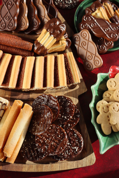 圣诞主题木质餐板上摆放着整齐的豪华比利时巧克力饼干、威化饼和酥饼的特写照片，背景是红色天鹅绒桌布和节日槲寄生餐具。图片下载