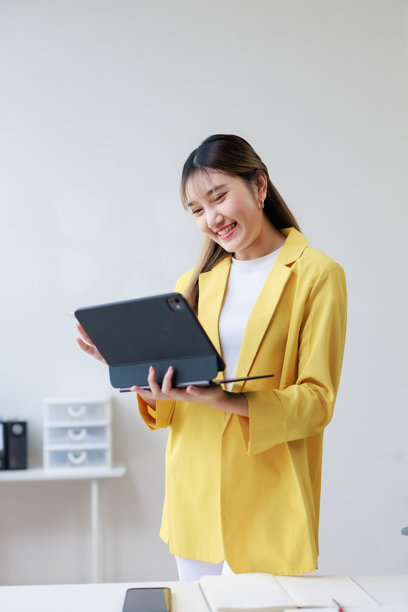 年轻的亚洲女商人，身穿鲜艳的黄色套装，自信地站在现代办公室里，她微笑着看着手中的数字平板电脑，似乎正在与屏幕互动。图片下载