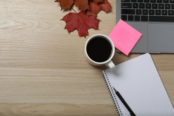 特写照片，俯视拍摄的笔记本电脑键盘、一杯饮品、贴纸、文件夹、回形针、笔记本和一支笔，以及黄色叶子，背景为孤立的棕色，留有空白空间。图片下载