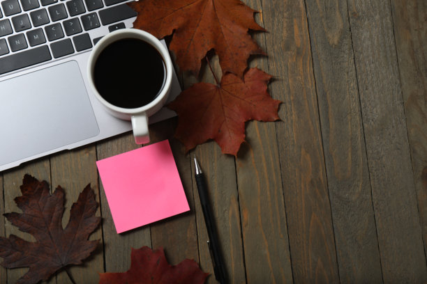 特写照片，笔记本电脑键盘、饮料杯、贴纸、文件夹、回形针、带笔的螺旋笔记本、黄叶平铺在棕色背景上，留有空白空间图片下载