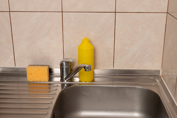 瓷砖墙背景下的厨房水槽，配有金属水龙头、黄色海绵和清洁剂塑料瓶。图片下载