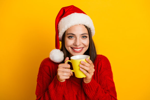 戴着圣诞帽的开心女孩穿着红色毛衣，手里拿着黄色马克杯，脸上带着灿烂的笑容，营造出完美的圣诞节日氛围，适合作为圣诞节素材照片。图片下载
