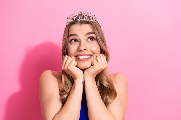 年轻貌美的女孩戴着皇冠，在粉色背景前微笑，非常适合时尚广告和生活方式的图库照片图片下载