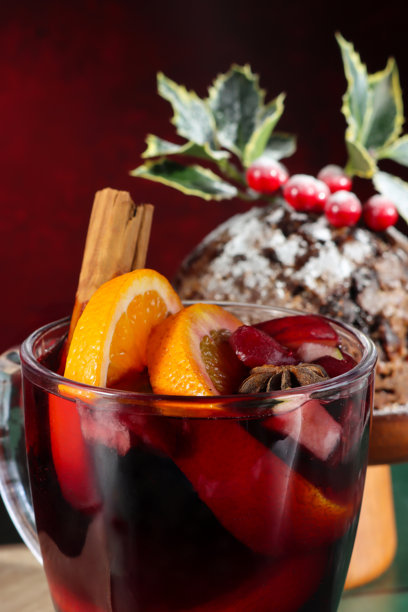 一张全画幅的格瓦斯酒图片，旁边放着一个木制蛋糕架，上面放着自制的圣诞布丁，装饰着冬青叶和红浆果，撒着糖粉，背景是绿色的表面和斑驳的红色，焦点在前景。图片下载