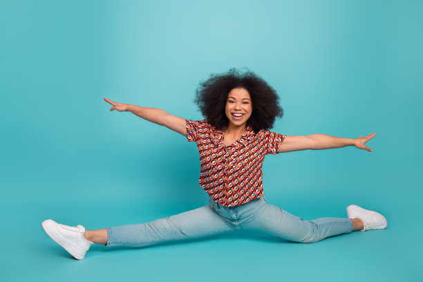 一位身穿色彩鲜艳印花衬衫的年轻女子在蓝色背景前做出完整的侧劈叉，面带微笑，构成一幅充满活力的生活方式、健身和时尚素材照片。图片下载