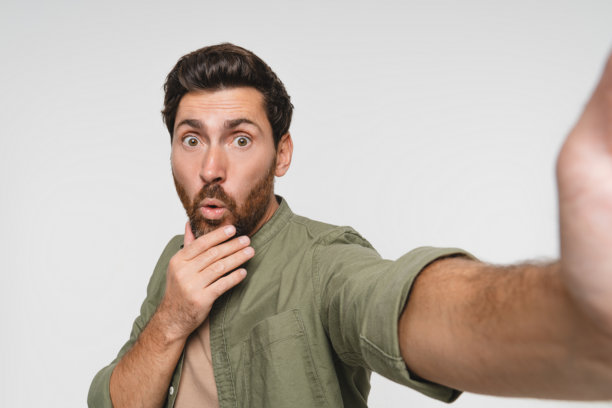 工作室白色背景下，一个男人自信地摆出有趣姿势自拍，适合发布或更新状态。这款应用、手机和照片展示了一位身着休闲装的网红，适合用作社交媒体头像。图片下载