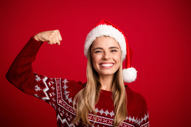 戴着圣诞帽、穿着红色圣诞毛衣的快乐女子，在节日营销照片中露出灿烂笑容，展示着她健壮的手臂。图片下载
