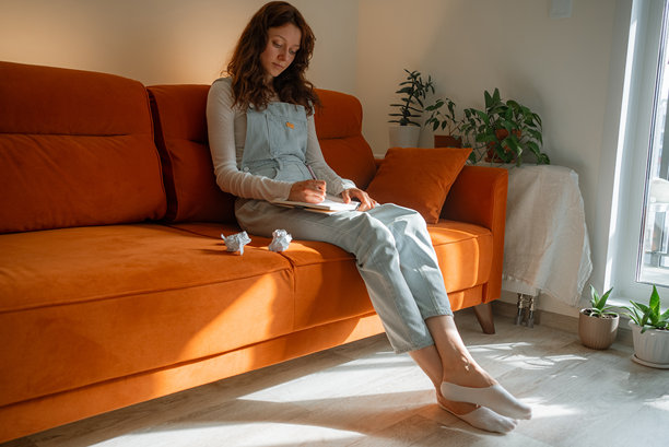 创意专业人士在明亮的室内工作空间中，坐在橙色沙发上，周围散落着揉皱的稿件和室内植物，正经历创作瓶颈。图片下载