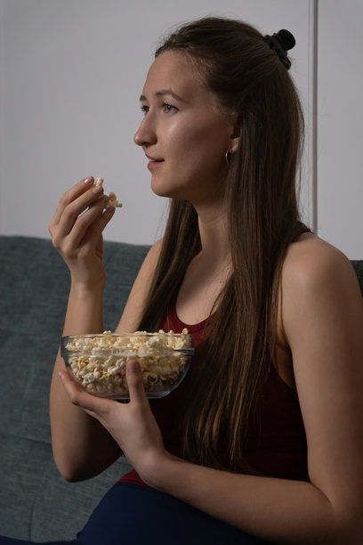 一位年轻女子微笑着坐在舒适的沙发上，手里拿着玻璃碗，在家一边看有趣的电影一边享受着吃爆米花。竖直照片图片下载