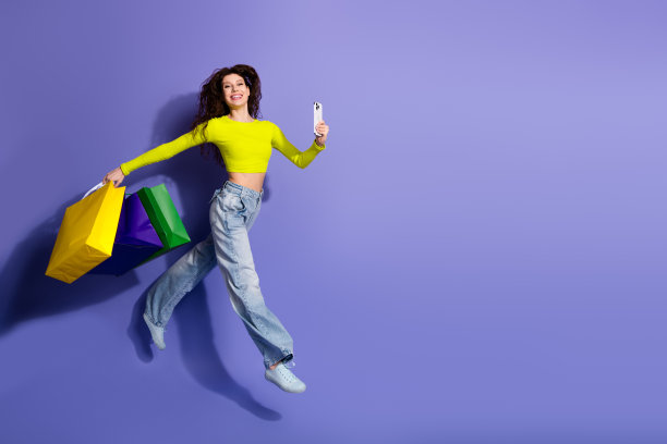 身穿亮黄色T恤的年轻女子拎着购物袋跳跃，同时对着紫色背景自拍，呈现时尚生活方式的图片图片下载