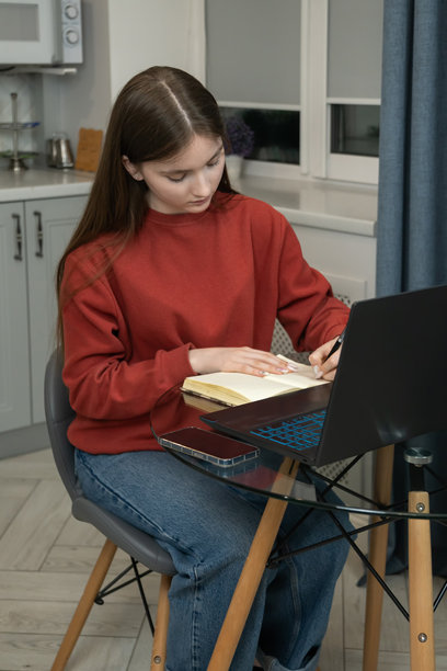 年轻女孩在厨房的笔记本电脑旁学习，在笔记本上写笔记，营造舒适高效的学习环境。竖直照片图片下载