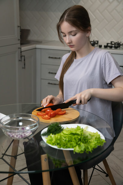 年轻女孩在现代厨房里，小心翼翼地在木质砧板上切新鲜番茄，旁边放着生菜，准备制作健康的餐食或沙拉。竖幅照片。图片下载