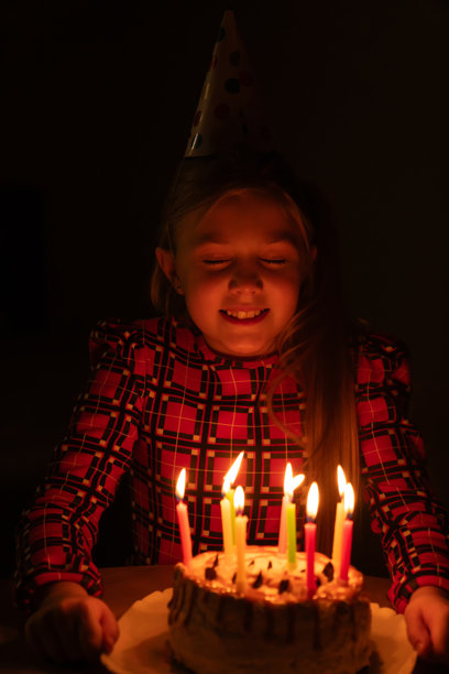 戴着派对帽的女孩闭着眼睛，在黑暗的庆祝活动中，对着生日蛋糕上明亮的生日蜡烛许下珍贵的愿望。竖屏照片图片下载