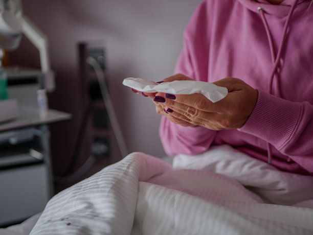 一名患者坐在病床上，手中拿着纸巾，在康复期间休息，一名穿着粉色连帽衫的女性出现在画面中。专业照片图片下载