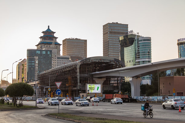 日落时分，阿斯塔纳的现代化城市景观，有新建的建筑和正在建设中的轻轨交通（LRT）线路。道路上有汽车和一名骑摩托车的人，建筑风格混搭，既有玻璃摩天大楼，也有东方风格的建筑。图片下载