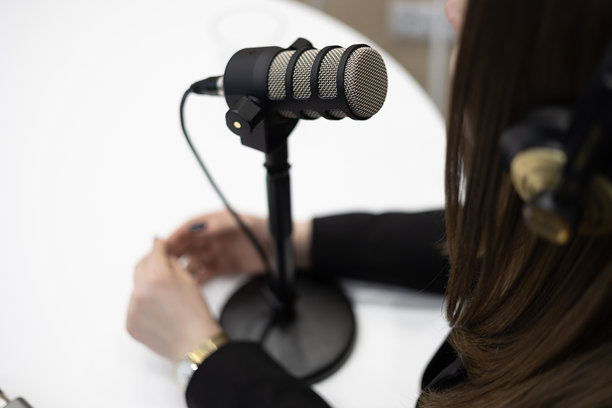 专业广播麦克风的特写和选择性对焦。一位女性播客或博主戴着耳机坐在桌前，正在说话或录制采访。图片下载