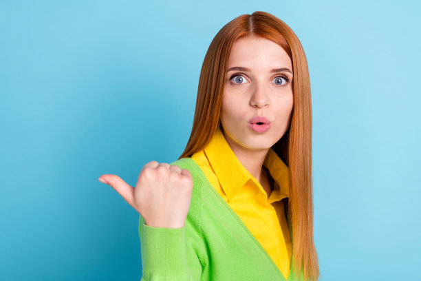 穿着黄色毛衣、在蓝色背景下展示惊叹表情的年轻女士的个人资料照片，手指指向空白处图片下载