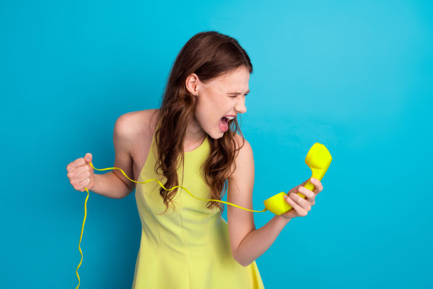 身穿时尚黄色连衣裙的年轻女子在明亮的蓝色背景前，对着复古电话热情地表达情感图片下载