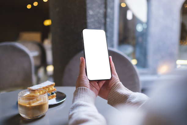 咖啡馆里，女子手持并使用手机，背景为空白的桌面屏幕的样机图图片下载