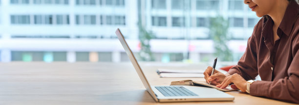 特写镜头：一位年轻的职业女性在办公室的办公桌前使用笔记本电脑，身后是城市景观。一位女经理或女学生在工作场所使用电脑进行工作或学习。横幅，可复制空间图片下载