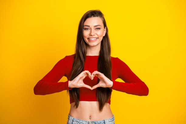 身穿红色露脐上衣的年轻女子对着明亮的黄色背景微笑，双手比心，适合生活方式和时尚素材图片图片下载