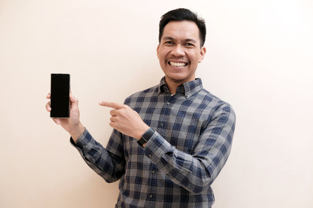 一位穿着法兰绒衬衫的东南亚男子，脸上带着开心的笑容，用手指着手机屏幕图片下载
