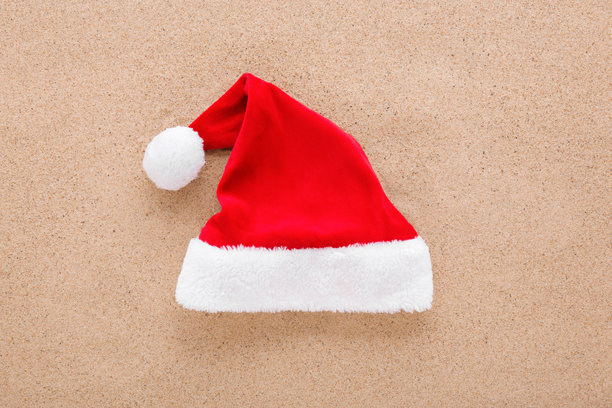 背景为干燥的浅棕色海沙，上面有一顶鲜红色的帽子，帽子边缘饰有白色毛皮。特写。圣诞节庆的服饰。俯视角度。图片下载
