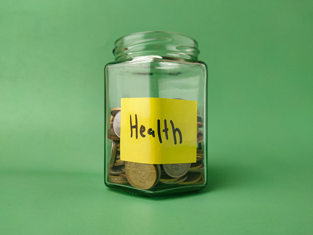 图片展示了一个装满硬币的玻璃罐，上面贴着一张黄色的标签，标签上手写着“健康”一词。图片下载