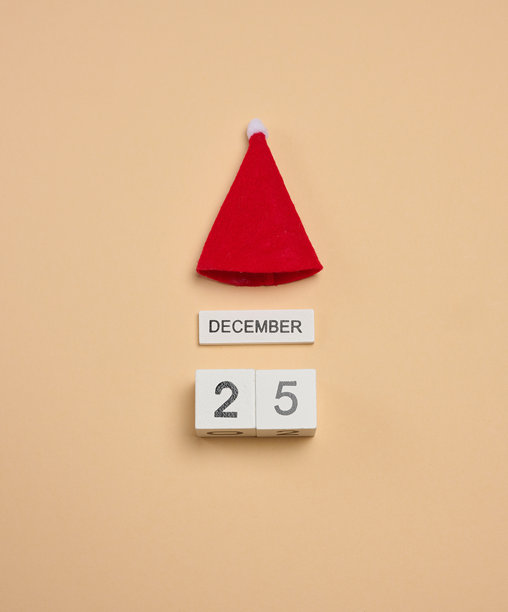 米色背景上，一个木制立方体日历显示日期为12月25日，上面还戴着一顶红色毛毡圣诞帽。图片下载