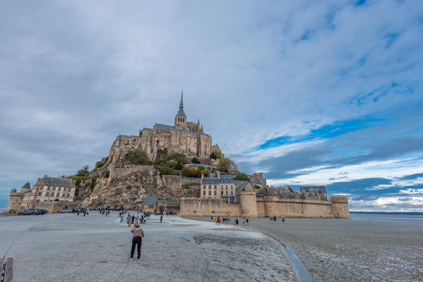 图片展示了宏伟的圣米歇尔山修道院，这是一座中世纪的修道院和堡垒，在部分多云的蓝色天空下，戏剧性地矗立在一座岩石山上。图片下载