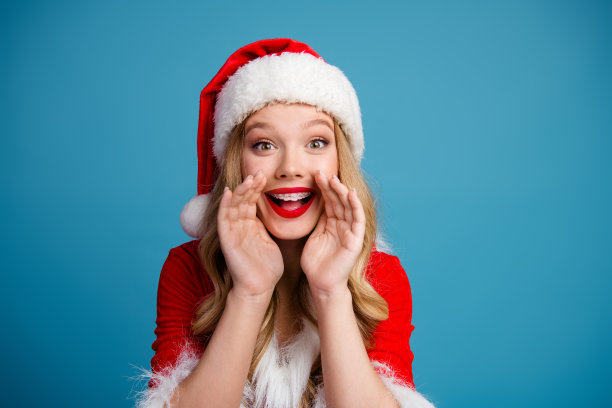 一位戴着圣诞帽、穿着红色连衣裙的微笑女士，在蓝色背景前拍摄的欢乐圣诞时尚照片图片下载