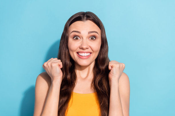 照片：女孩在柔和的蓝色彩色背景前，喜悦地笑着，做出胜利者的姿势。图片下载