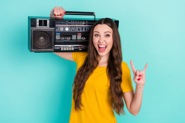 身穿黄色T恤的年轻酷女孩，一手拿着复古收音机，一手做出摇滚手势，背景是鲜艳的蓝绿色图片下载
