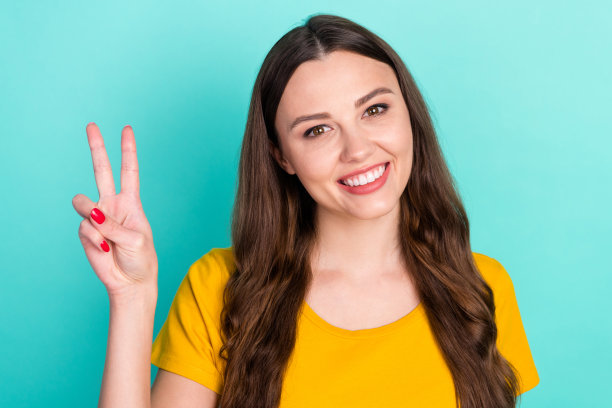 身穿黄色衣服、比着V字手势、面带微笑的迷人可爱年轻女子照片，背景为蓝绿色图片下载