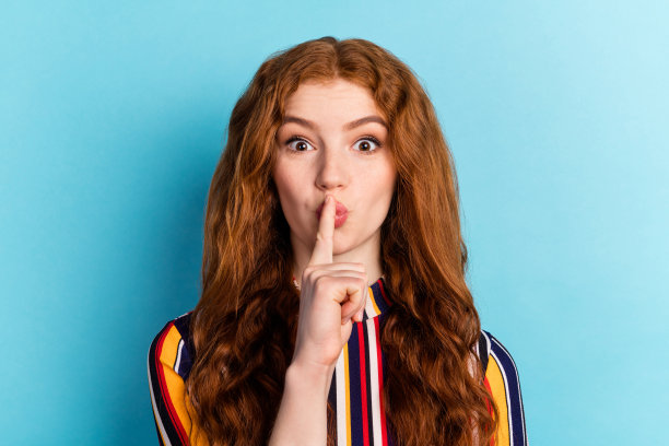 身着彩色衬衫的年轻女子，拥有疯狂的红色发型，手指放在嘴唇上，在蓝色背景前摆出姿势的特写照片图片下载