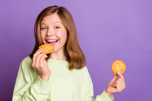 照片：一个年轻的学龄前女孩，带着快乐积极的笑容，吃着美味的饼干甜点，孤立在紫色背景上图片下载