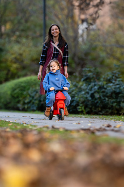 蹒跚学步的孩子在小路上骑着玩具摩托车，母亲在一旁陪伴图片下载