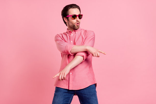 照片：一个时髦、年轻、快乐、开朗的男人，手指着舞者，音乐爱好者，孤立在粉彩粉色背景上图片下载