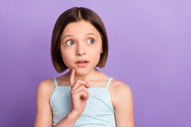 照片：惊讶震惊的年轻小女孩，手指托着下巴，目光望向空处，孤立在紫色背景上图片下载