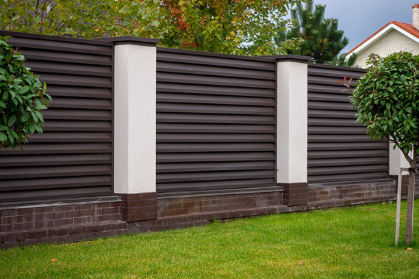 现代深棕色复合材料围栏，配有白色柱子和砖砌基座，环绕着绿色草坪。用于说明隐私屏风、物业边界和现代住宅入口设计。真实照片图片下载