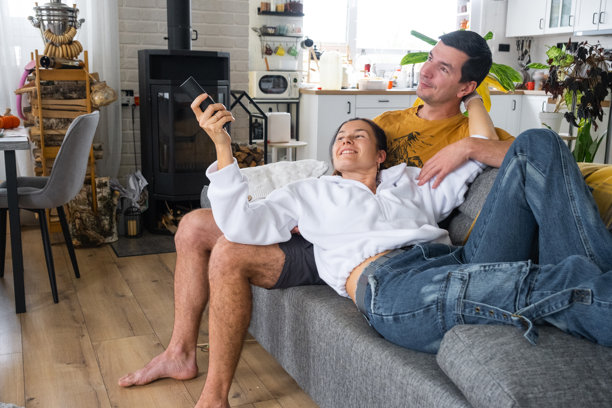 一对情侣在家中的白色内景里坐在沙发上看电视。居家休闲、娱乐、爱情、家庭、约会图片下载