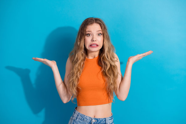 一位穿着橙色上衣、表情困惑的迷人年轻女子，耸肩站在蓝色背景前，用于时尚生活方式图库照片图片下载