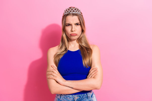 头戴皇冠的年轻女子身穿蓝色上衣，双手交叉，站在粉色背景前，显得自信而时尚，非常适合时尚生活风格的图库照片图片下载