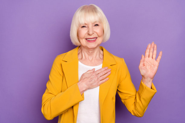 照片：一位乐观、漂亮的短发老太太，穿着黄色夹克，在紫色背景前祈祷图片下载