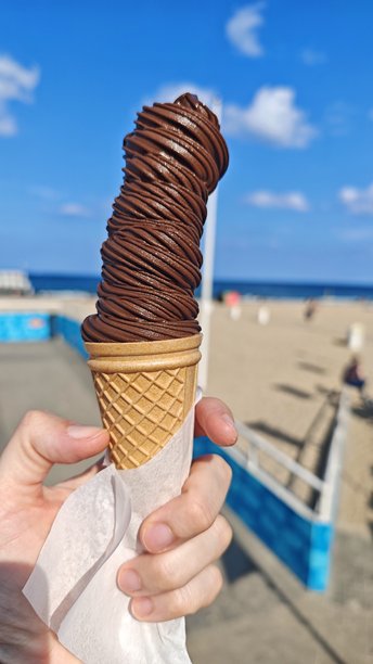 一只手拿着装在华夫脆筒里的高旋巧克力冰淇淋，背景是阳光明媚的海滩。这张图片传达了享受、夏日甜点和无忧无虑的休闲时光。图片下载
