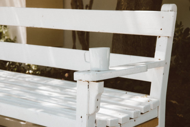 柔和的户外光线下，一个白色咖啡杯放在复古白色木凳的扶手上，极简的视角，宁静的庭院氛围，带有质朴的纹理，适合创意视觉使用的放松与生活方式概念图片下载