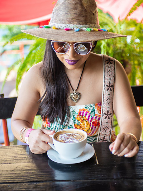 戴着帽子和太阳镜的女士正在享用一杯带有精美拿铁拉花艺术设计的咖啡。图片下载