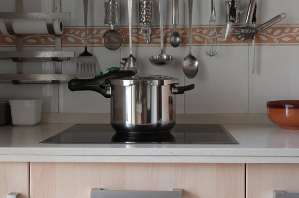 现代厨房里，时尚的黑色电磁炉上放着一个不锈钢压力锅，旁边悬挂着金属厨具，明亮简约的台面暗示着高效的家庭烹饪。图片下载