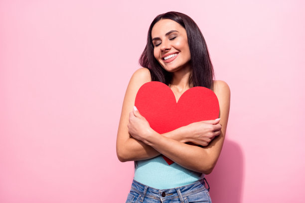 年轻女子手抱红色爱心明信片，在柔和的粉色背景前露出喜悦的笑容，照片肖像图片下载