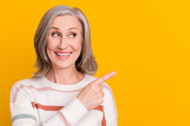 照片：一位身穿白色毛衣、面带微笑、竖起手指指向空处的闪亮甜美退休女性，背景为孤立的黄色。图片下载